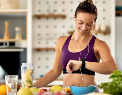 Športnica v športni opravi stoji v kuhinji, obkrožena z zdravo hrano, kot so sadje in smoothie. Gleda na uro na svoji zapestnici, kar simbolizira načrtovanje prehrane pred ali po vadbi